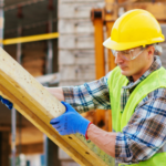 Construction worker in protective gear lifting and positioning a large wooden beam at an active jobsite.