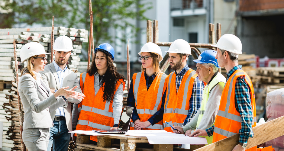 Team of construction professionals, including designers and site crew, gathered around project plans on a jobsite, illustrating the importance of coordinated teamwork in building projects.