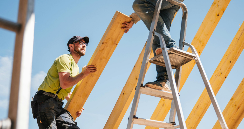 Construction workers installing wooden beams, with one worker lifting a board and another standing on a ladder at a building site.